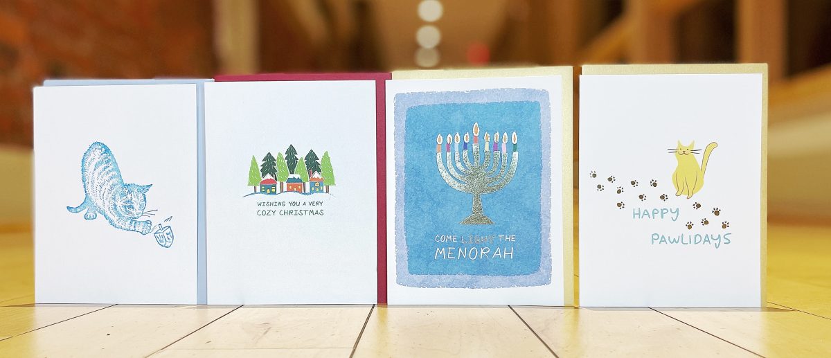 Four holiday cards in a row, depicting a cat playing with a dreidel, a snowy Christmas village with the words 'Wishing you a very cozy Christmas,' a lit menorah on a blue background with the words 'Come light the menorah,' and a smiling cat surrounded by brown paw prints, with the words, 'Happy pawlidays.'